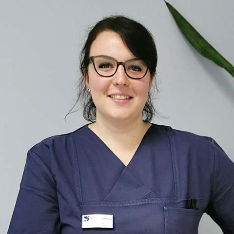 Veterinärassistenz in dunkler medizinischer Kleidung steht in einem Innenraum neben einer Pflanze.